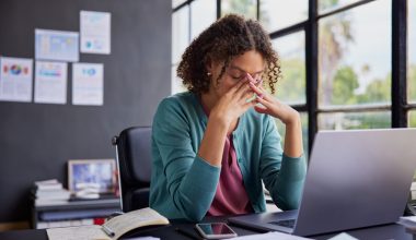 Photo: adult woman with curly hair wearing a green cardigan over a pink top sits at a desk, eyes closed and fingers pressed to her face, appearing stressed while working on a laptop with papers, notebook, and phone nearby.