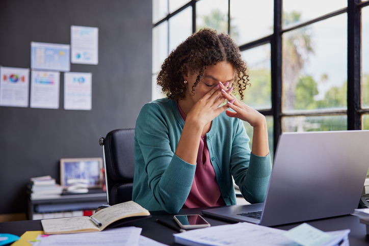 Photo: adult woman with curly hair wearing a green cardigan over a pink top sits at a desk, eyes closed and fingers pressed to her face, appearing stressed while working on a laptop with papers, notebook, and phone nearby.