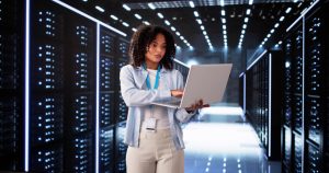 Photo: adult woman with curly dark hair wearing a light blue shirt, ID lanyard, and beige pants, using a laptop while standing in a data center aisle lined with illuminated server racks.