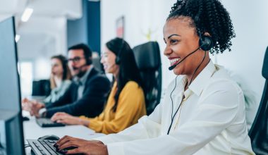 Photo of customer service representative wearing a white button-down shirt and headset, typing at a computer workstation in a modern call center, with three additional headset-wearing colleagues and monitors visible in the background.