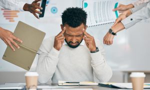 Photo: Person in a light gray sweater seated at a desk, pressing fingers to temples while multiple coworkers’ hands extend a smartphone, folder, printed chart, and pointing gestures toward the person; office setting with documents, notebook, calculator, and coffee cups on the desk.