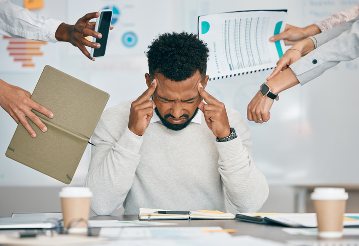 Photo: Person in a light gray sweater seated at a desk, pressing fingers to temples while multiple coworkers’ hands extend a smartphone, folder, printed chart, and pointing gestures toward the person; office setting with documents, notebook, calculator, and coffee cups on the desk.